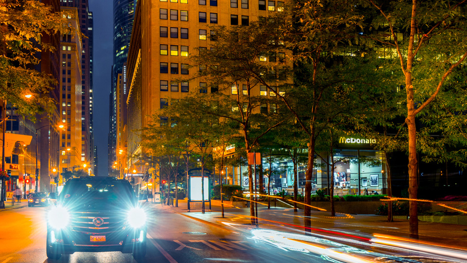 Ein Auto fährt nachts in einer Großstadt. Am rechten Fahrbahnrand stehen Bäume.